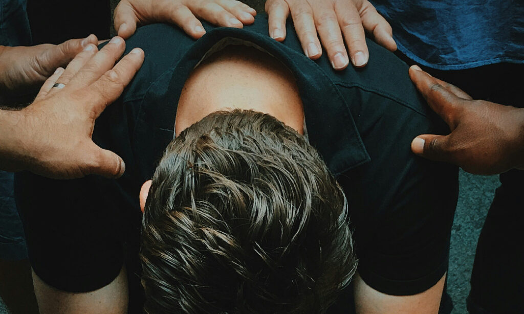 Fotografía desde arriba de un joven con la cabeza hacia abajo mientras unas manos se posan sobre su espalda en señal de apoyo, utilizada para el artículo del blog que trata sobre los talleres de bullying y las vivencias como facilitadoras y facilitadores, de Javi Castillo Rubio, Laura Torrella y Carles Porrini