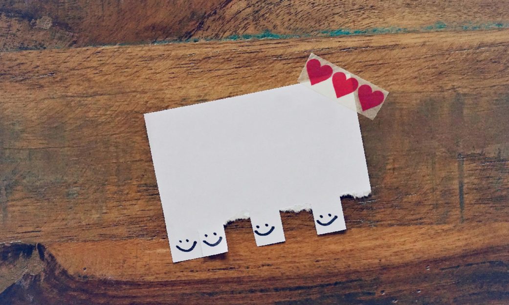 Fotografía de un anuncio en blanco, con caritas sonrientes para coger, con algunas que ya han sido arrancadas, y un sticker de corazones en el extremo superior derecho, fotografía utilizada para la actividad sobre el cuidado, la empatía y la propia consciencia de nuestras emociones