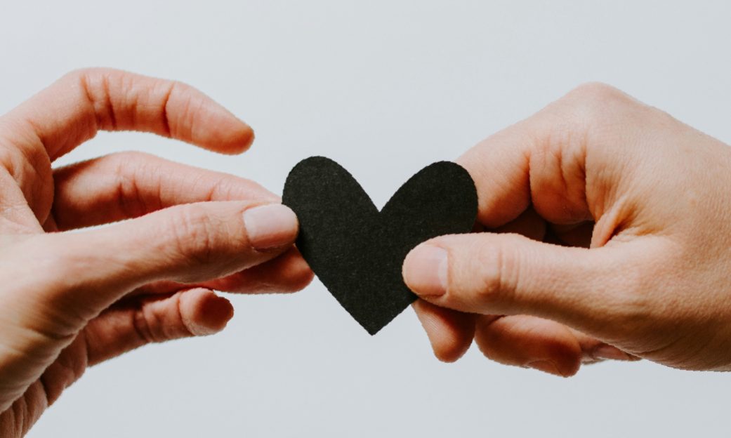 Fotografía de dos manos sosteniendo con los dedos un corazón de cartulina, cada mano de un extremo, utilizada para la actividad sobre las emociones y relaciones dentro del aula para ofrecer propuestas de mejoras