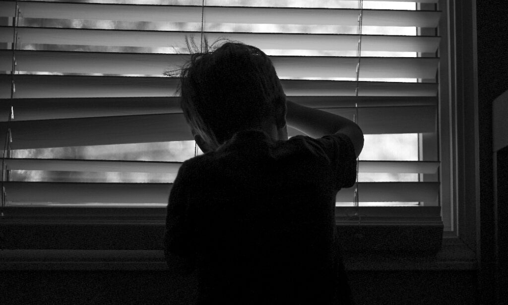 Fotografía de un niño de espaldas mirando por una ventana con la persiana cerrada en blanco y negro, y reflejando angustia, utilizada para el pódcaste donde se explica la participación de Equip Seer y PDA Bullying en la creación del manifiesto frente a las Violencias Sexuales Infantiles (VSI)